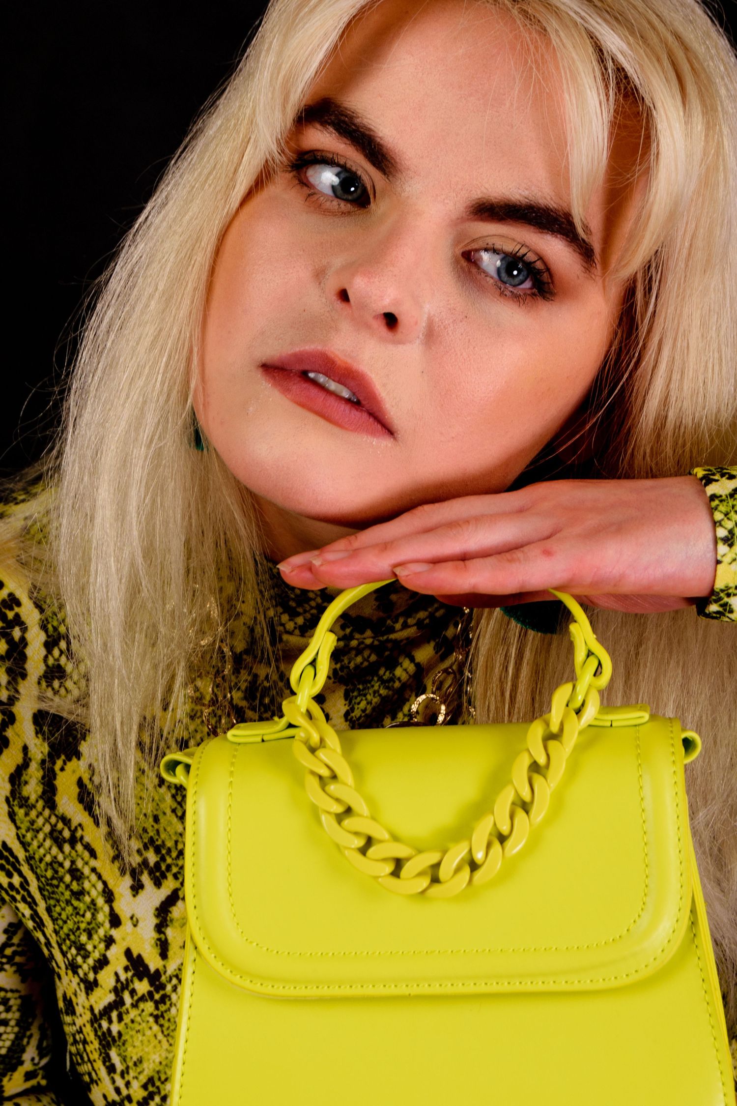 Blonde woman in yellow snakeskin top holding a neon yellow chain-handle handbag against a black background.