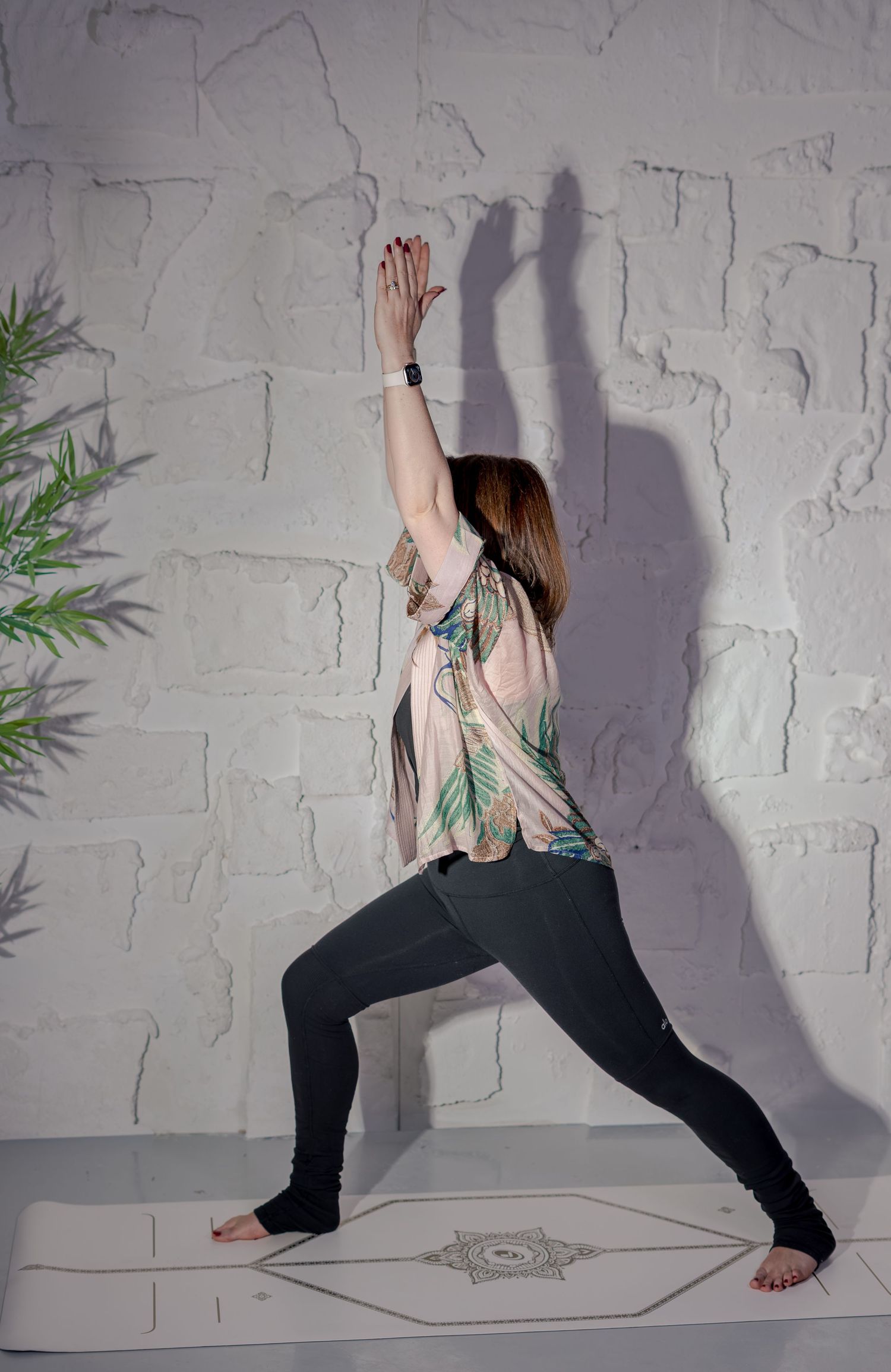 Woman practicing yoga warrior pose on mat against white brick wall, wearing floral top and black leggings.