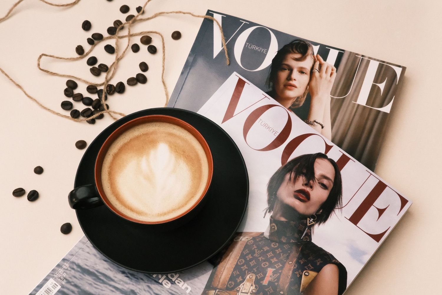 A black cup of latte art beside two Vogue magazines and scattered coffee beans on a beige surface.