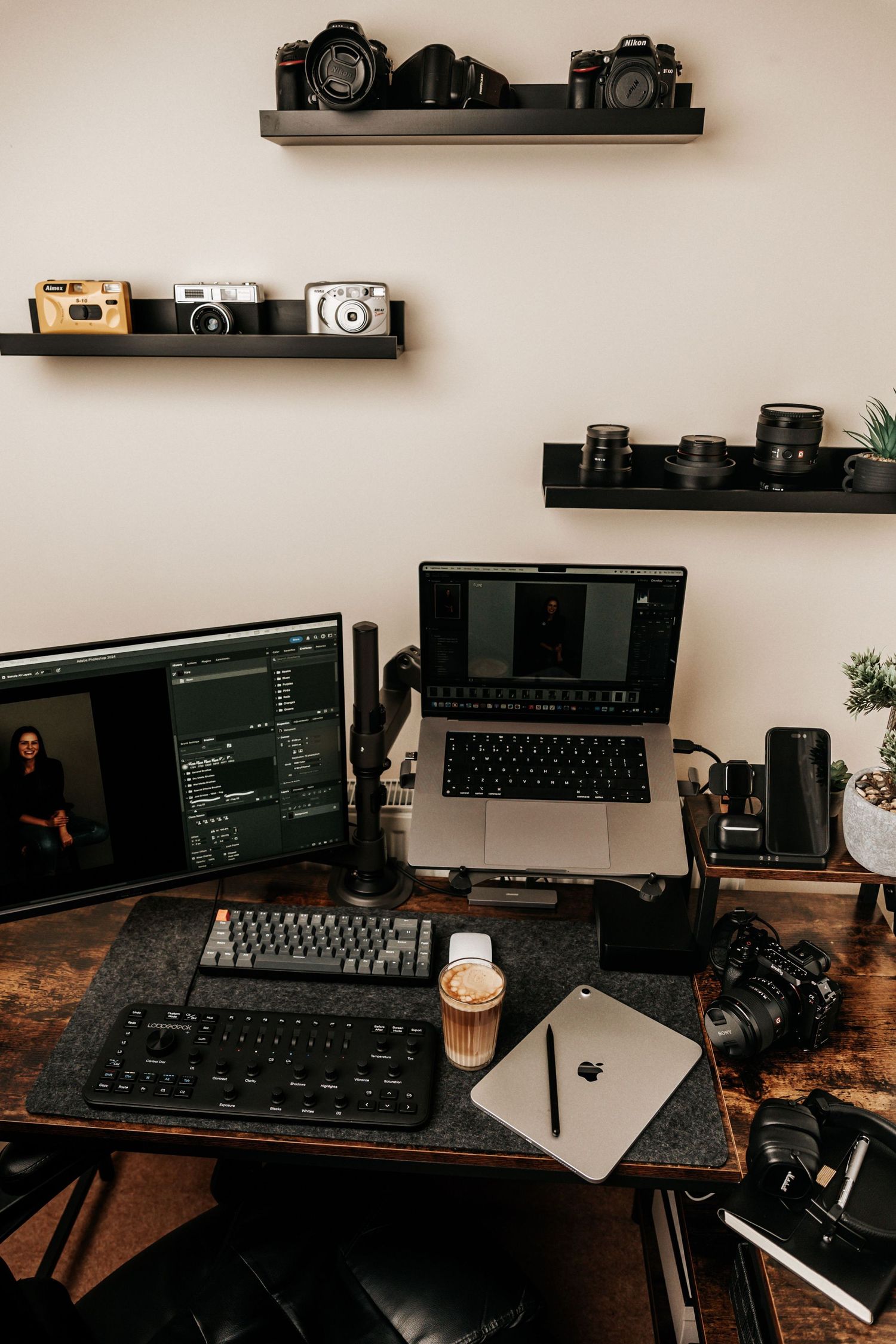 Photographer's desk setup with MacBook Pro, dual monitors, keyboard, cameras, and vintage camera collection on wall shelves.