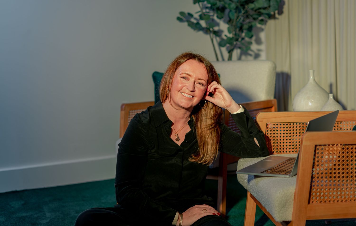 Woman smiling in black outfit seated in cane chair against teal wall with plant decor.
