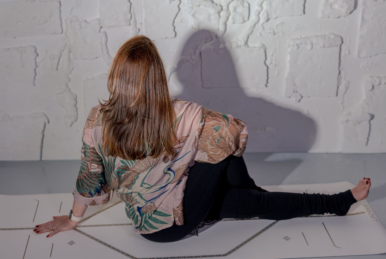 Woman with red hair in floral jacket sits on white floor, back to camera, casting shadow on textured wall.
