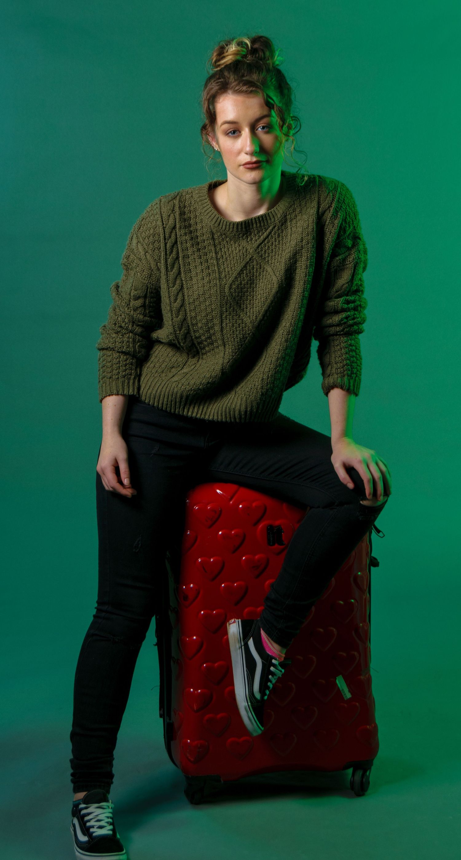 Casual portrait with green sweater while sitting on red patterned luggage against green background.