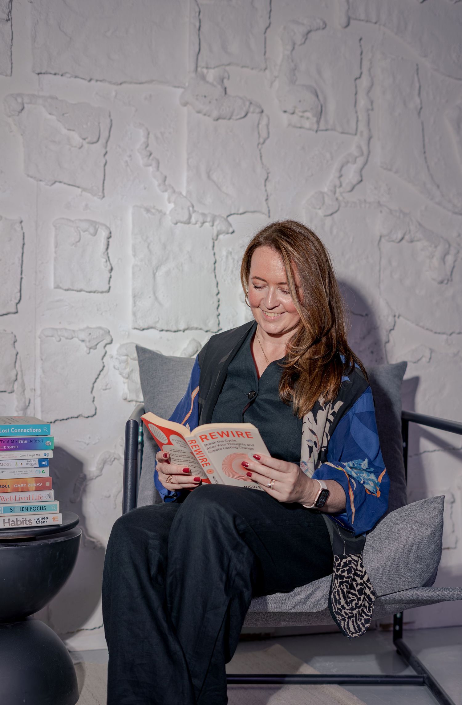 Woman smiling while reading a book titled Rewire, seated in a modern chair against a textured white wall.
