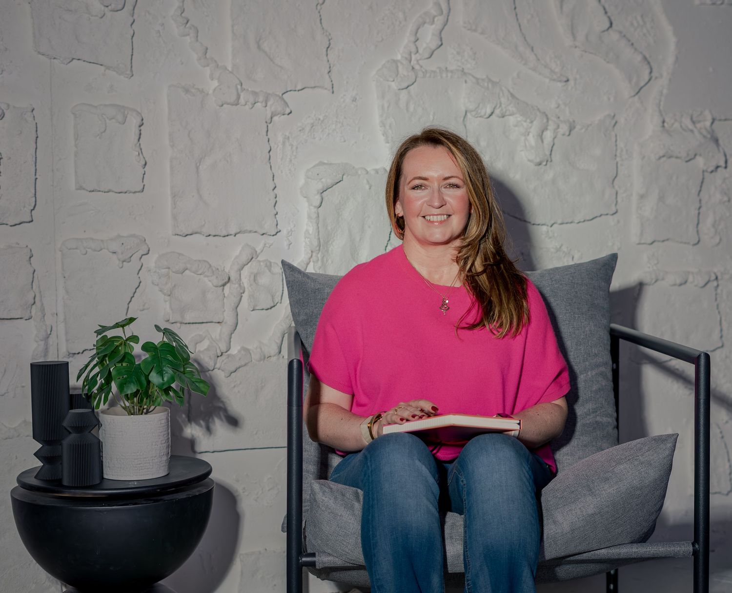 Smiling woman in pink top sitting in gray chair holding a book beside a plant against white wall.