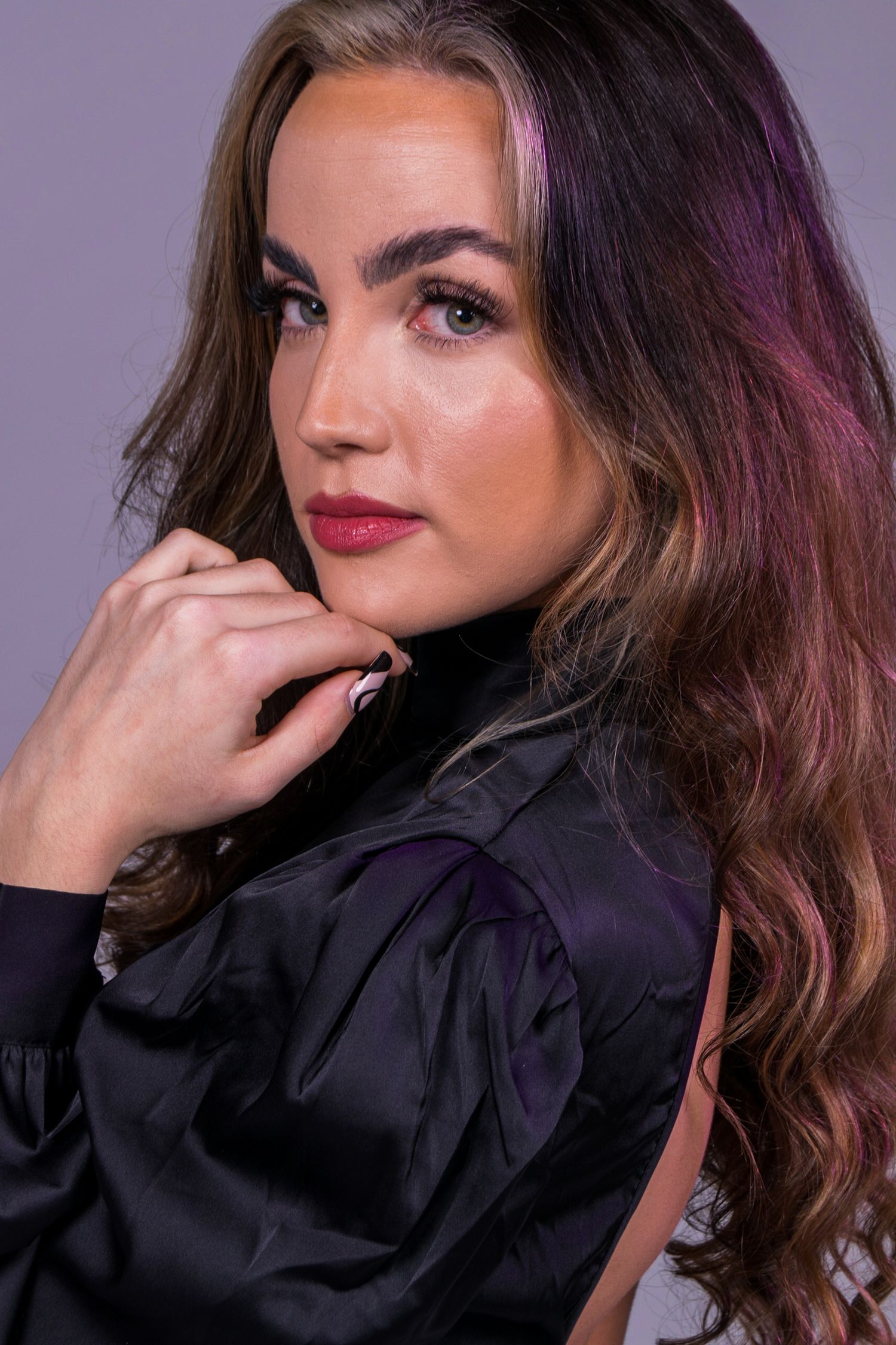 A woman with long brown hair in a black satin top poses confidently with her hand near her chin against a purple backdrop.
