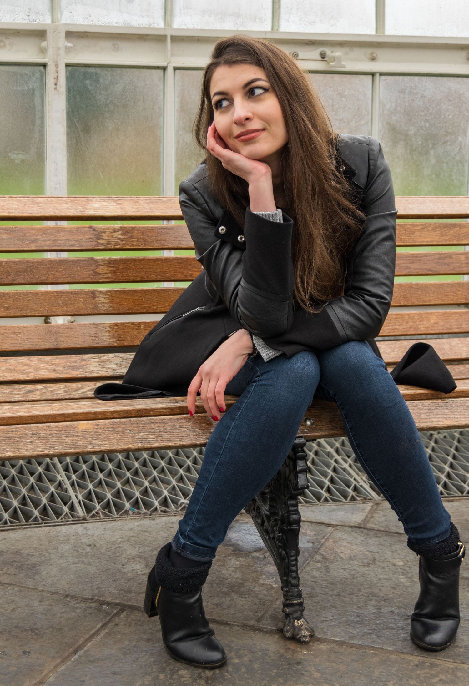 Young woman in black leather jacket and jeans sitting thoughtfully on a wooden bench outdoors.