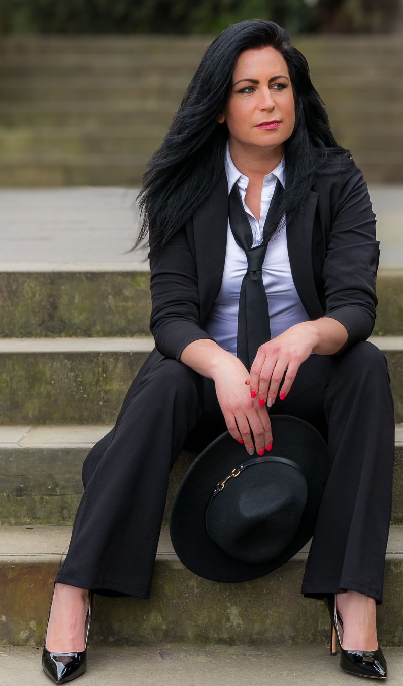 Woman in black suit and tie sitting on steps holding a black hat with red nails and patent heels.