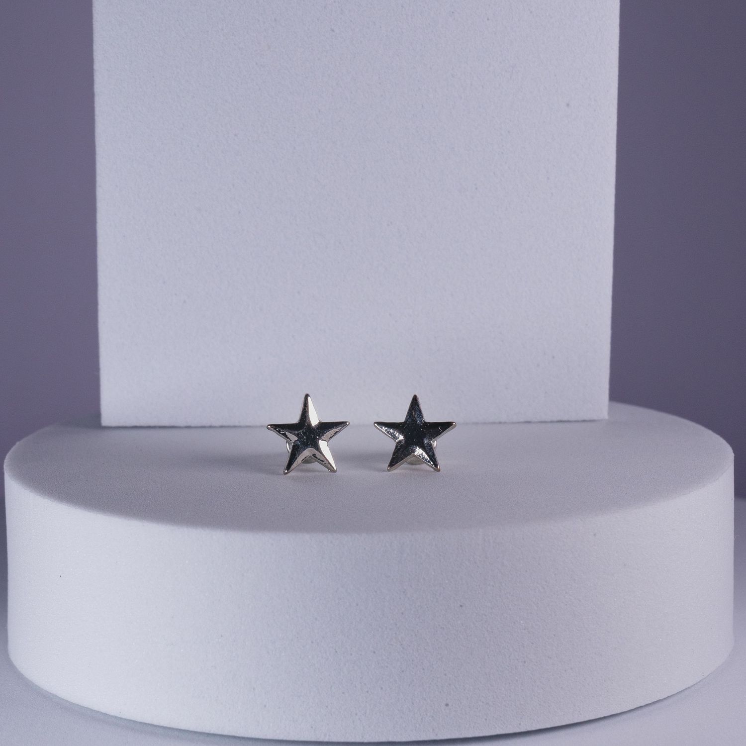 Pair of small silver star stud earrings displayed on a white cylindrical pedestal against a light background.
