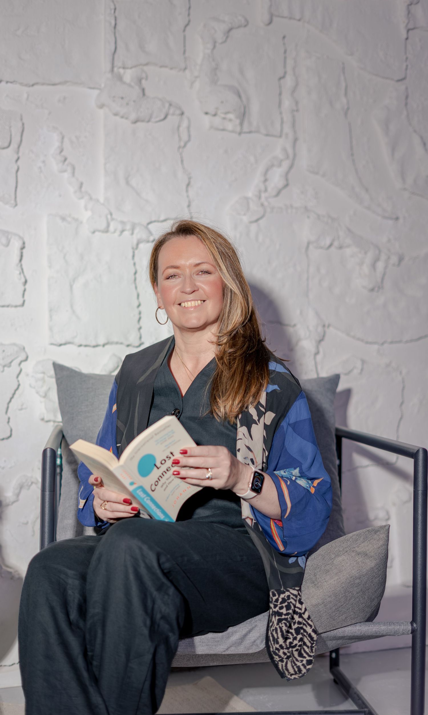Smiling woman sitting in a chair holding a book, wearing a blue floral blouse, with a textured white wall behind her.