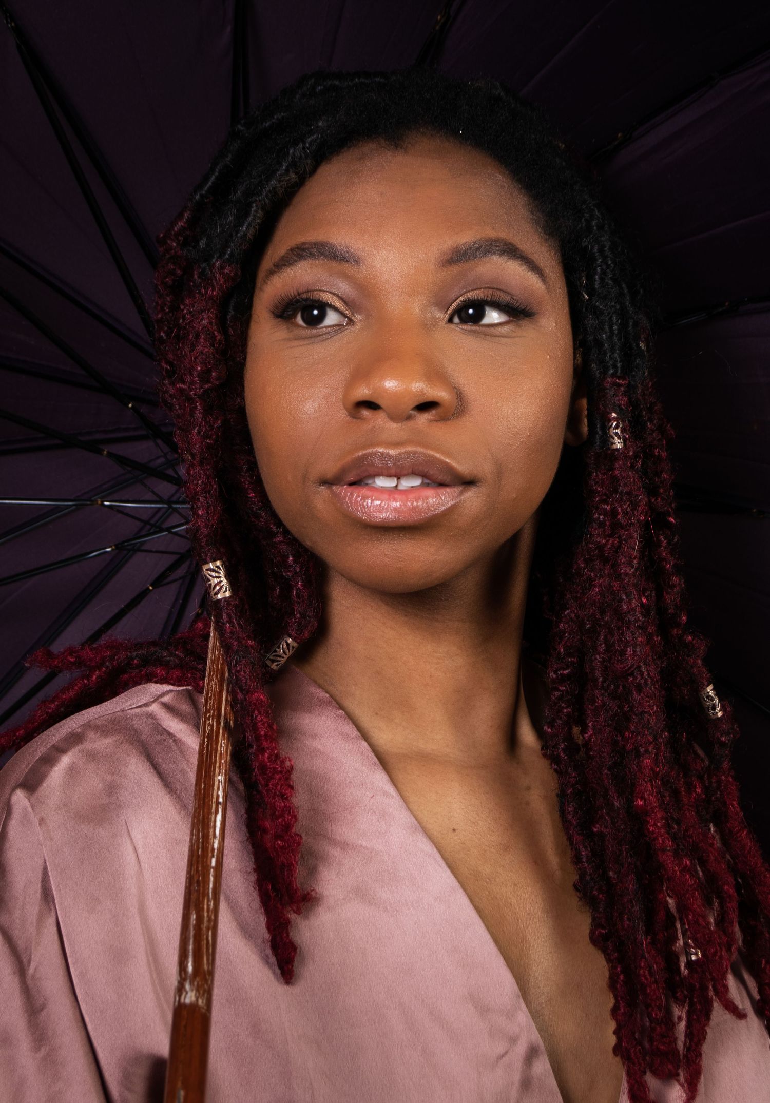 Young woman with red-tipped locs holds a dark umbrella, wearing a mauve top, studio portrait.