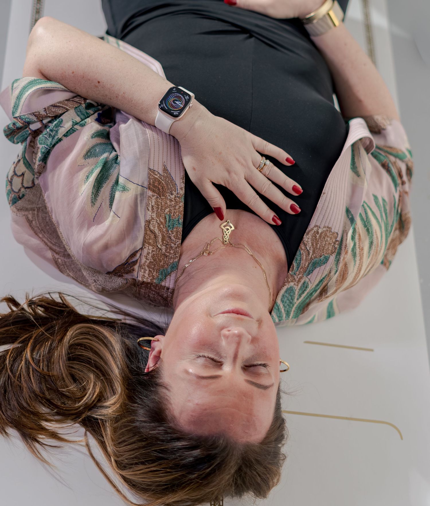 Pregnant woman lying down, wearing floral kimono, black dress, Apple Watch, and red nails, cradling her baby bump.