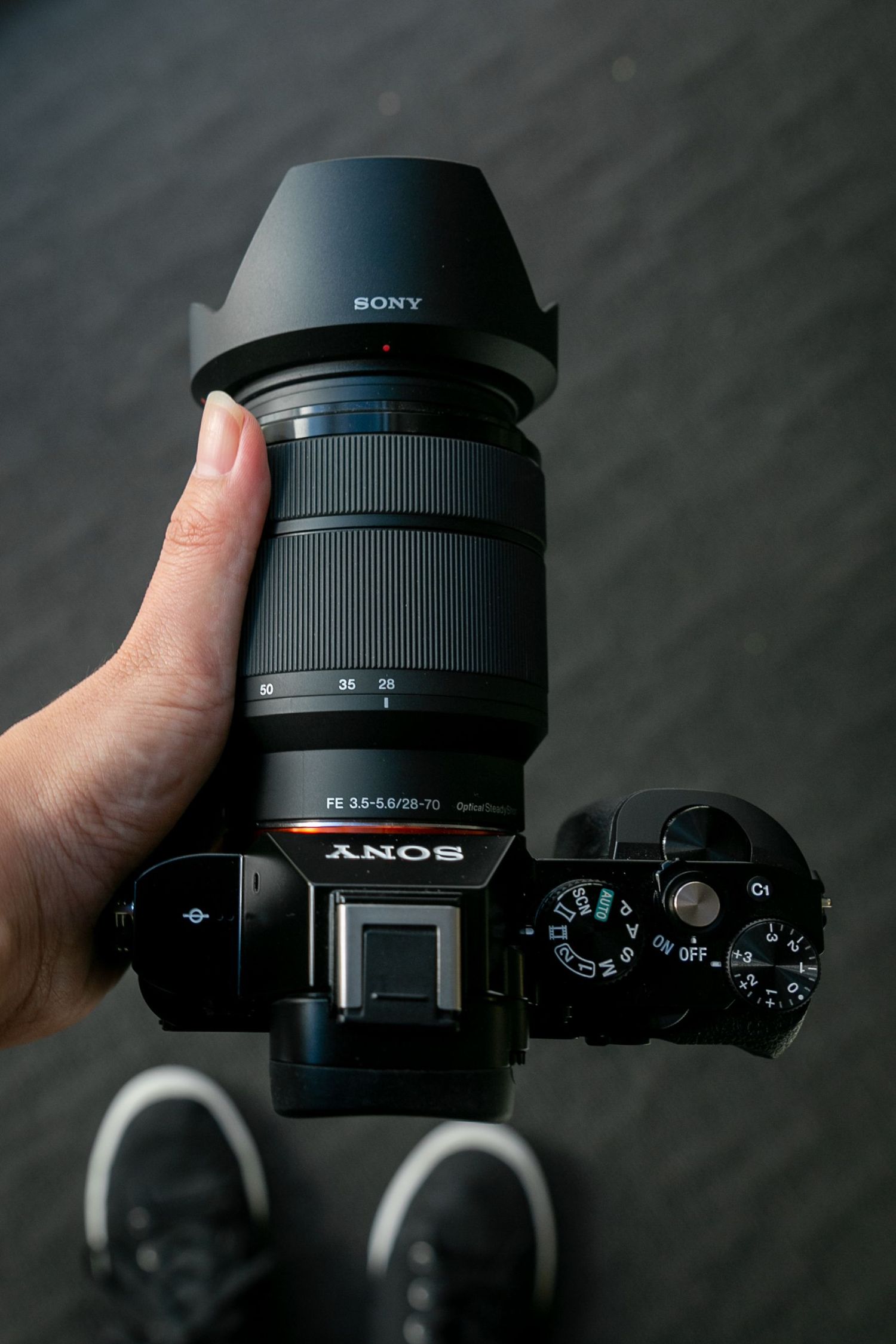 Hand holding a Sony mirrorless camera with FE 28-70mm zoom lens and petal lens hood, viewed from above.