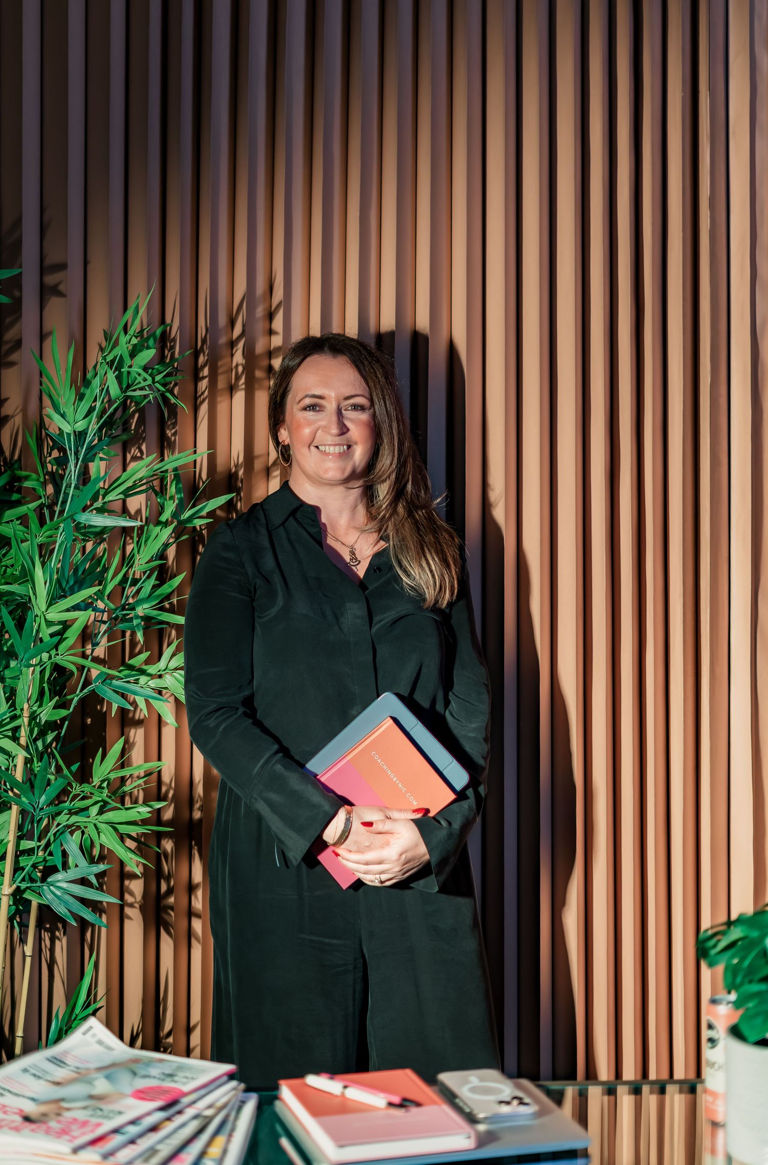 Woman in dark green outfit holding colorful notebooks, smiling beside a plant against a wooden slat wall.