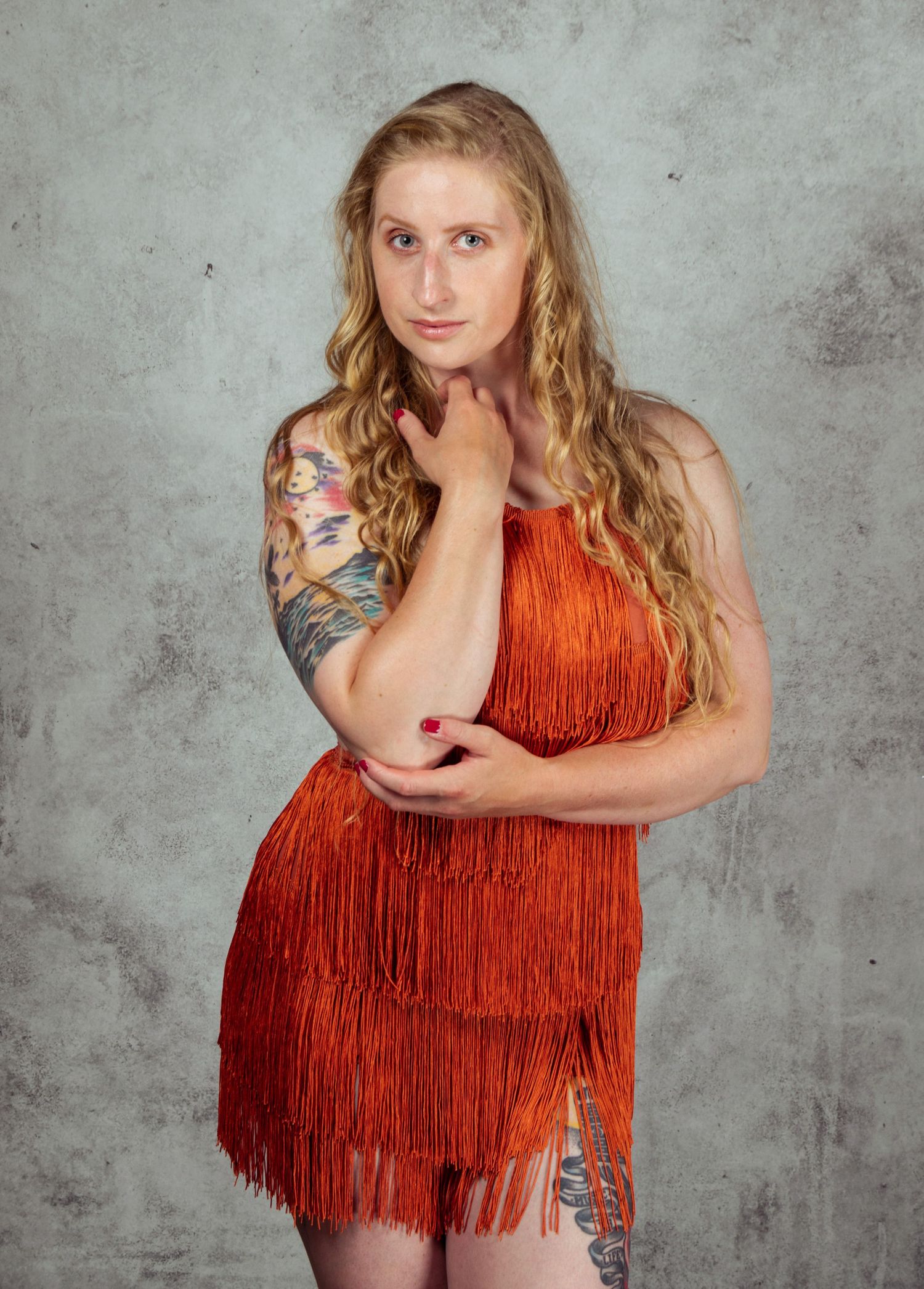Model in orange fringe top with tattoos poses against textured grey wall.