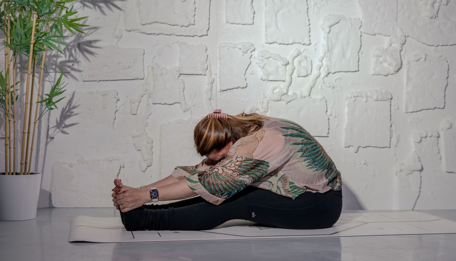 Woman with tattoos performing a seated forward bend yoga pose on a mat against a white brick wall.