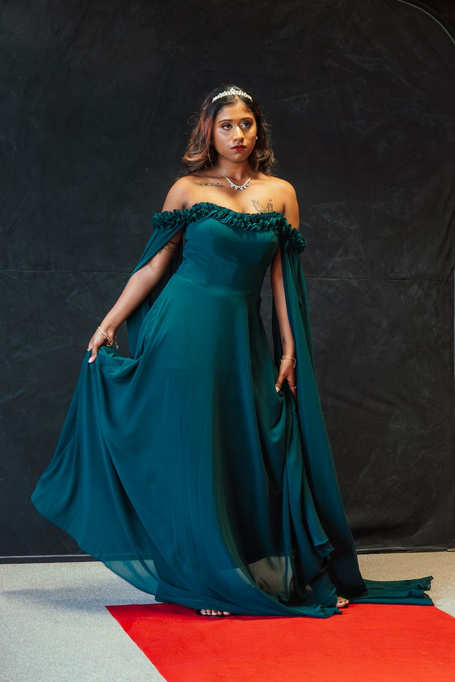 Woman in elegant emerald green off-shoulder gown with cape detail and tiara posing on red carpet.