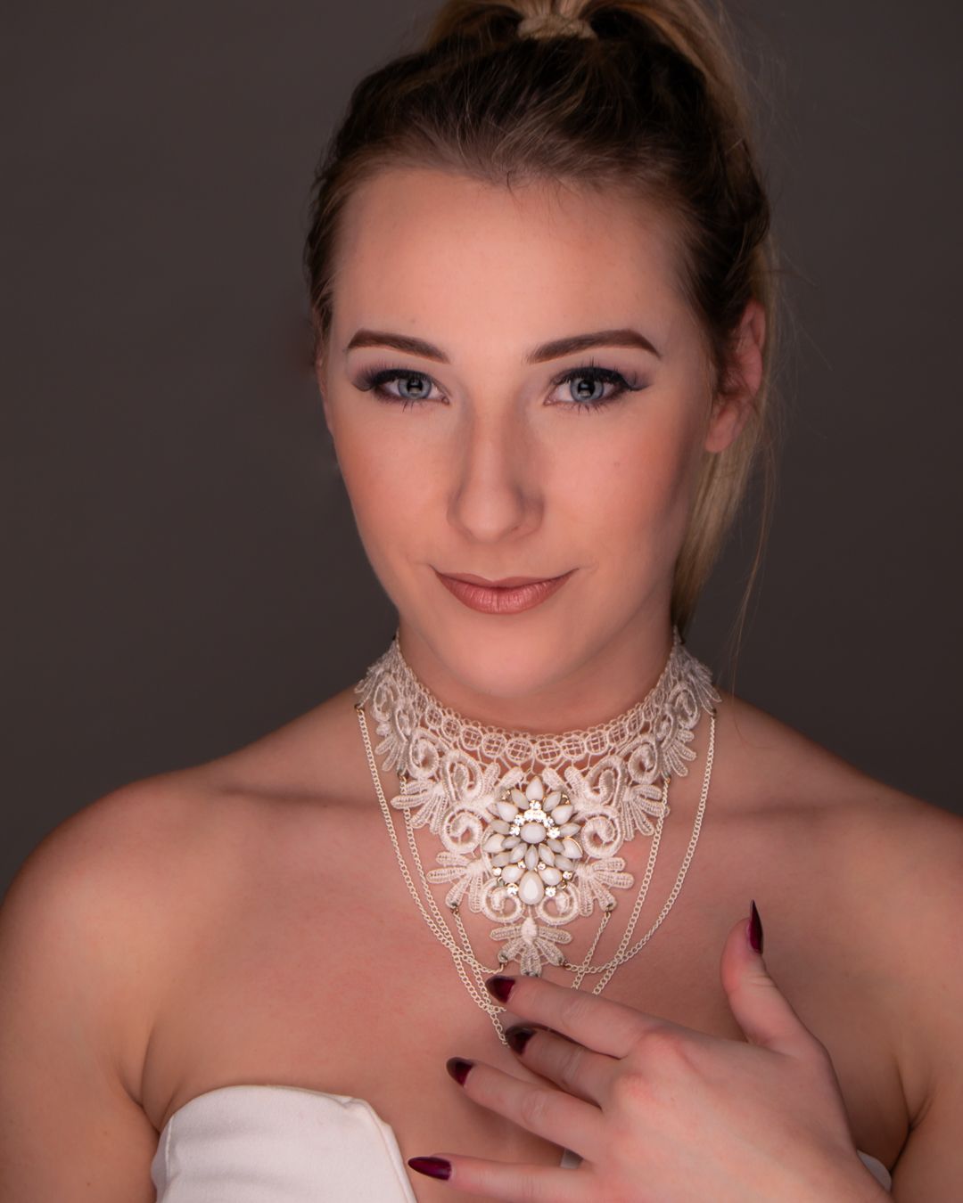 Young woman with blonde updo wearing elegant white lace choker necklace, posing against dark gray background.