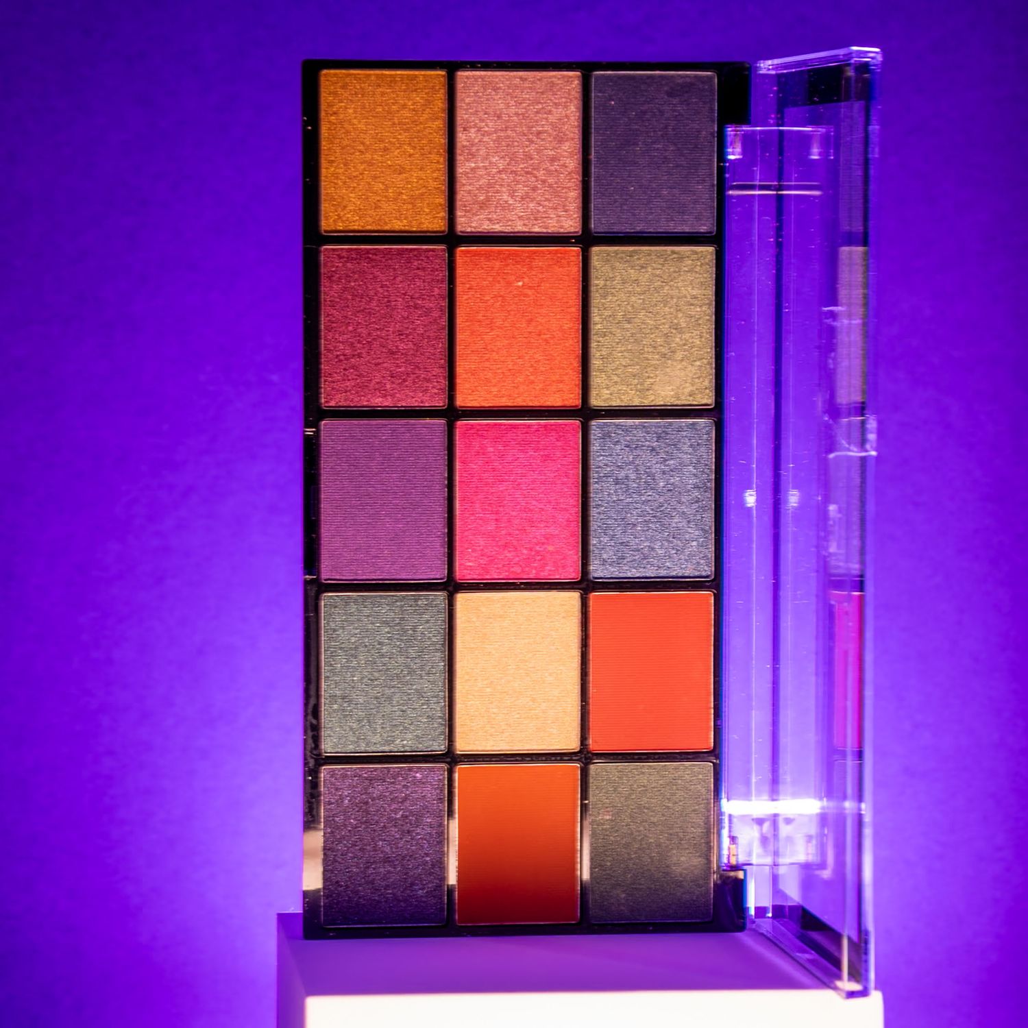Vibrant eyeshadow palette with 15 shades in orange, pink, purple, and gold tones displayed against a purple background.
