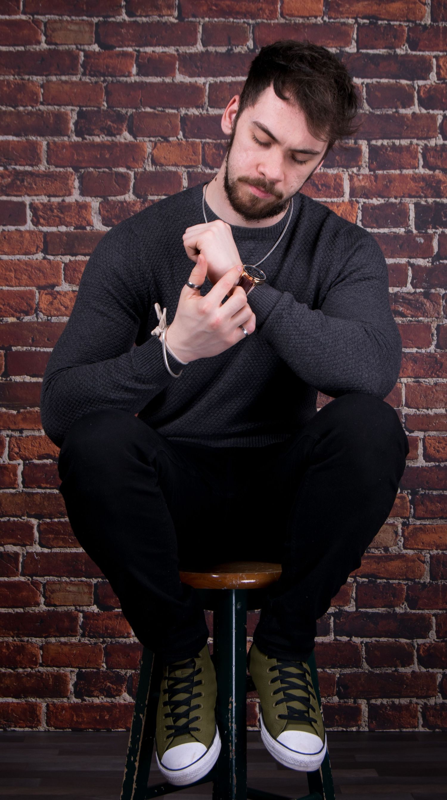 Young bearded man sitting on stool against brick wall, wearing gray sweater, black pants, and olive green sneakers.