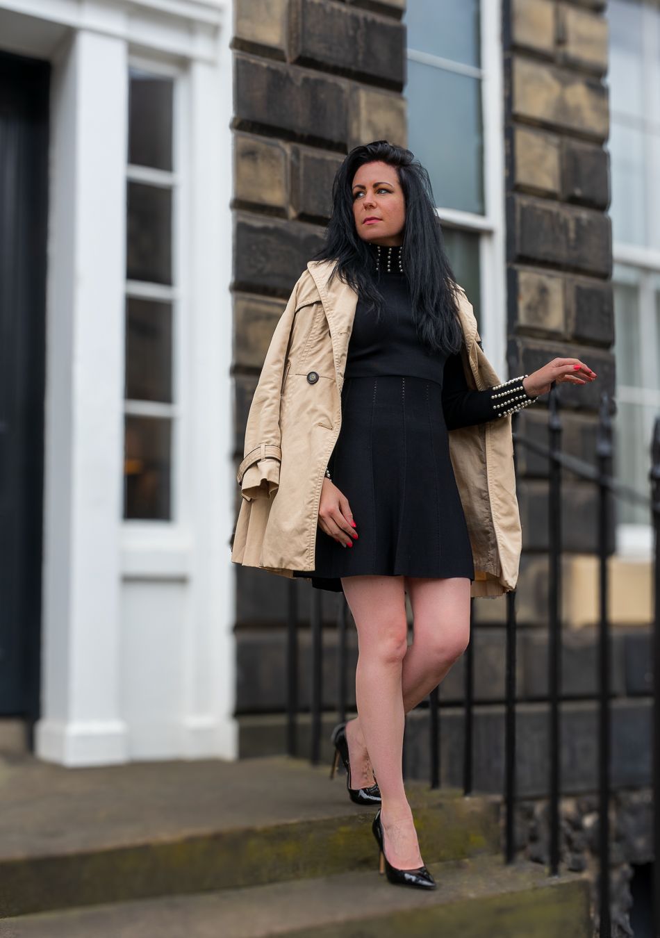 Woman in black dress and beige trench coat posing on stone steps outside a classic building wearing black heels.