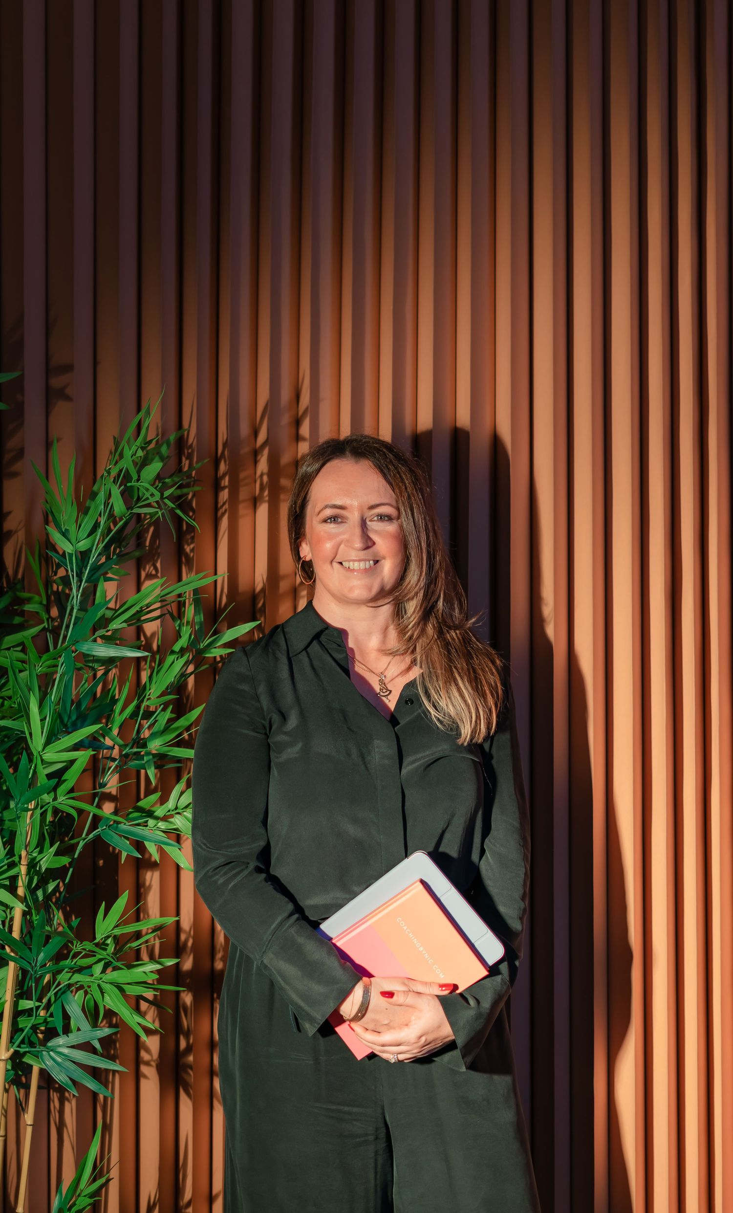 Professional woman in green outfit holding notebooks, smiling against wooden slat wall with plant.