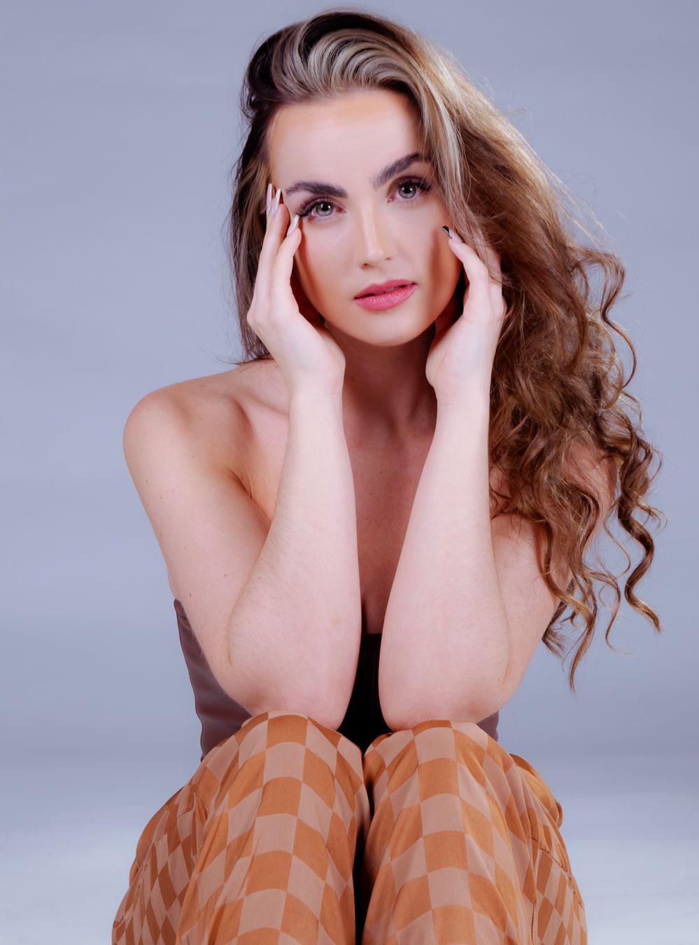 Young woman with long curly hair poses seated, hands framing face, wearing checkered orange pants against gray background.