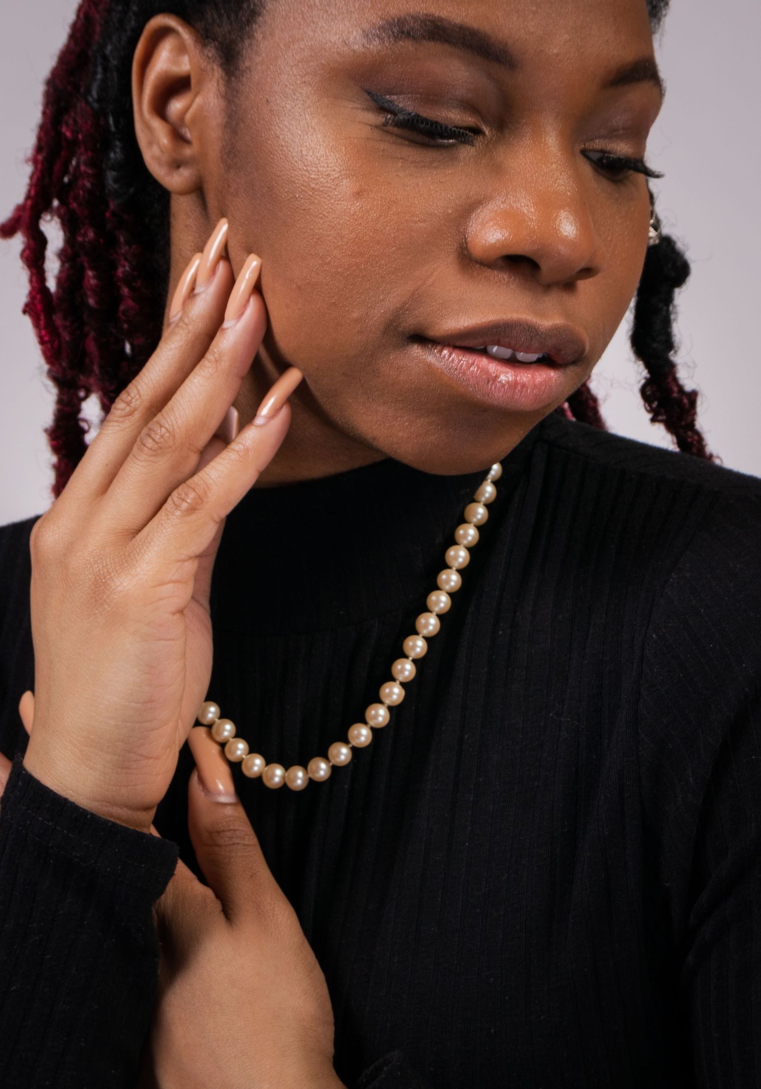 Woman wearing a pearl necklace and bracelet set, dressed in black, with red braids and neutral nails.