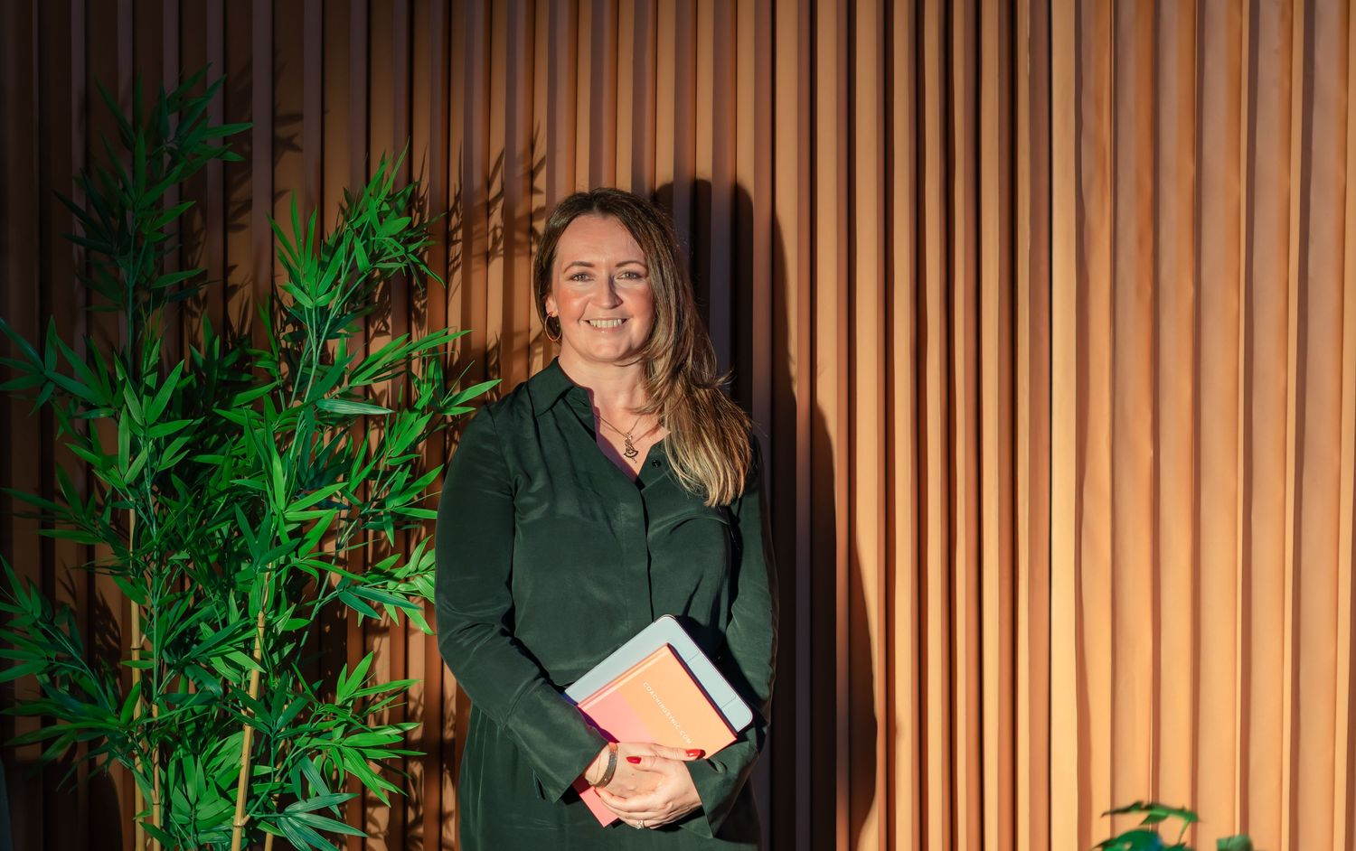 Woman in green outfit holding books stands smiling beside bamboo plant against wooden slat wall.