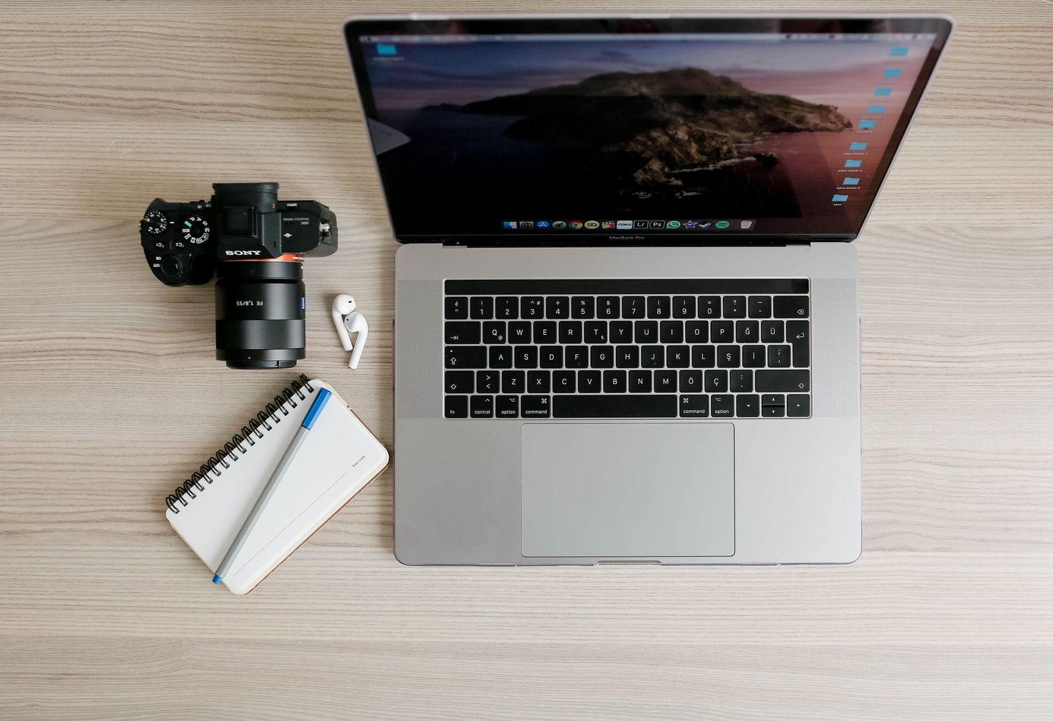 MacBook Pro laptop, DSLR camera, spiral notebook, and wireless earbuds arranged on a wooden desk.