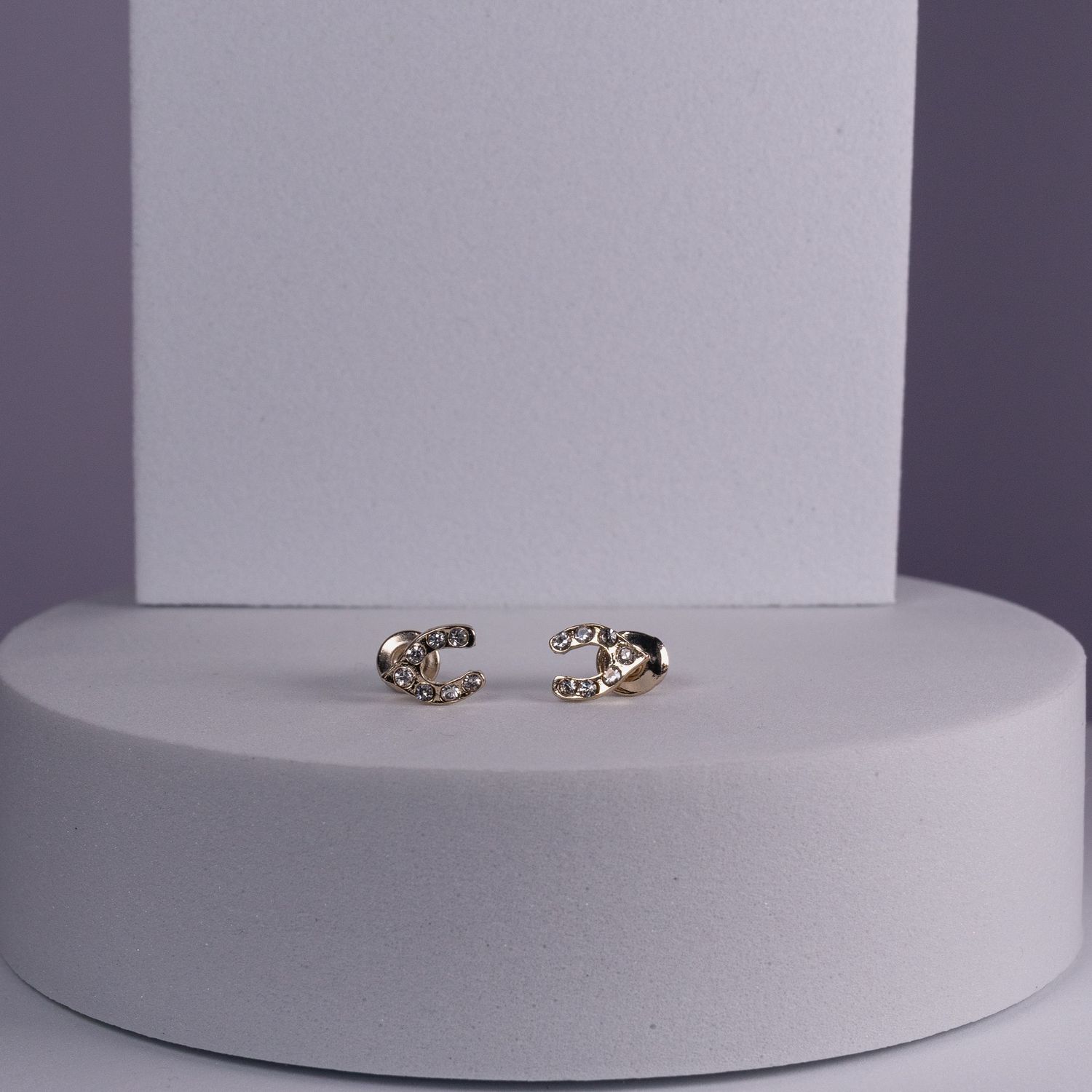 Gold-tone crystal embellished letter C stud earrings displayed on a white pedestal against a gray background.