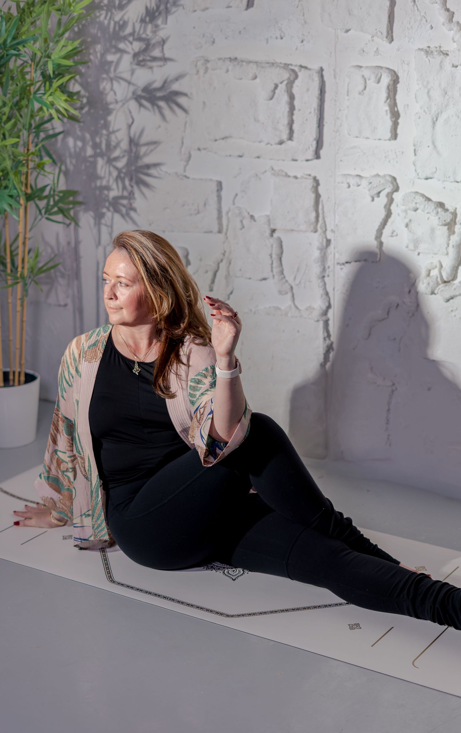 Woman in black yoga outfit with floral wrap sits on yoga mat against white brick wall with bamboo plant.
