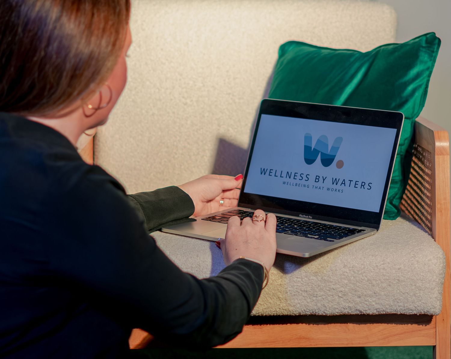 Woman using laptop displaying Wellness by Waters logo while seated in a cozy chair with green pillow.