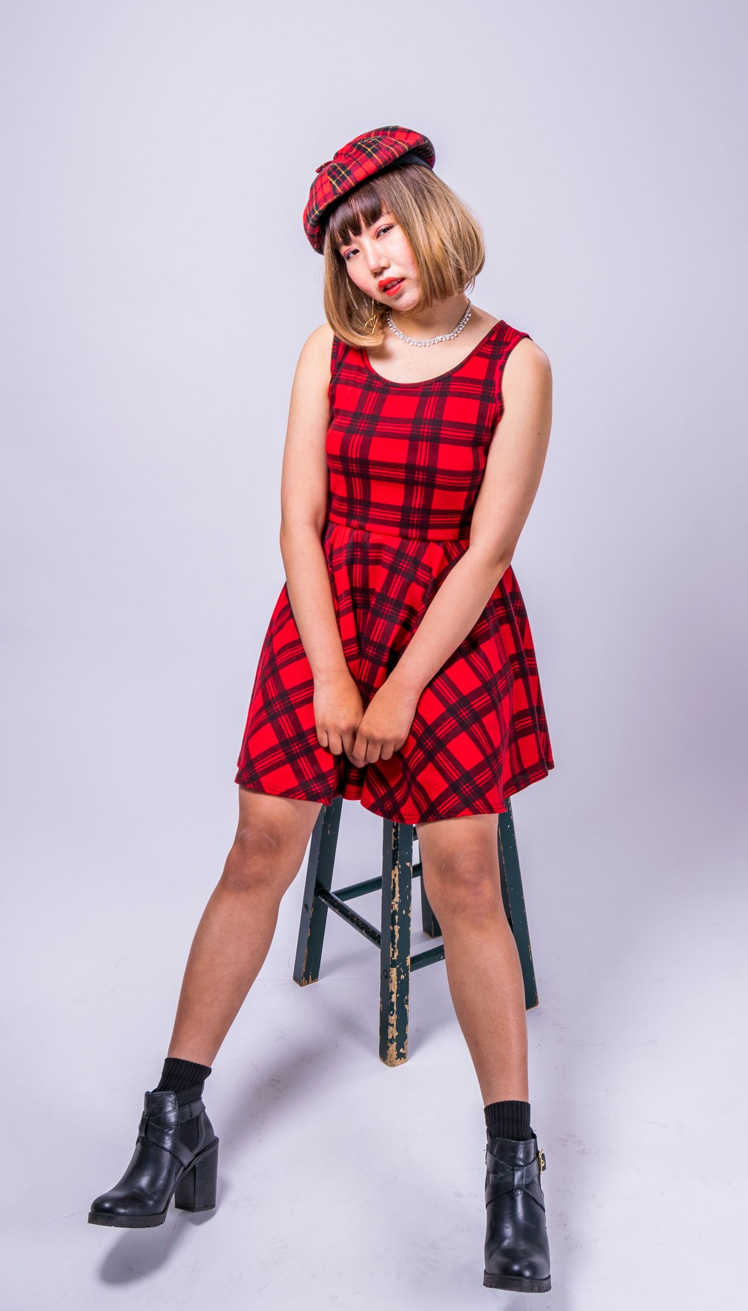 Playful pose in red plaid dress and matching beret with black boots against light background.