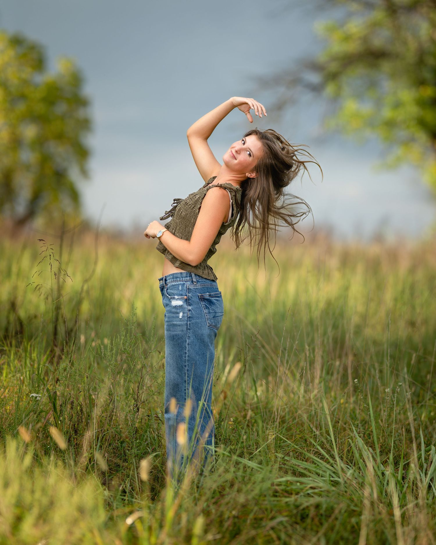 Young woman in jeans and green top poses with hair flowing in the wind on a cloudy day. Jen Welker, Kansas City Senior Portraits
