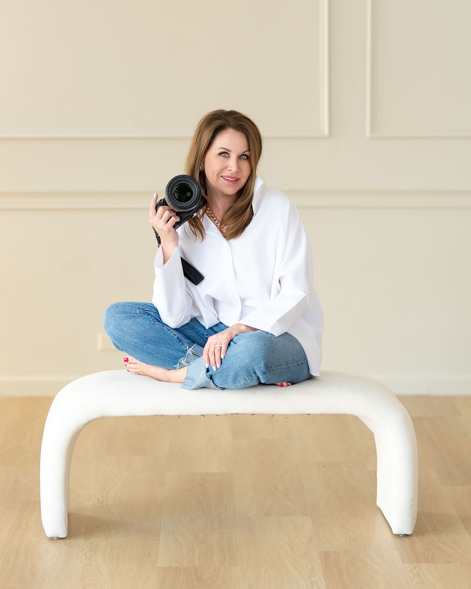 Woman photographer sitting cross-legged on white bench holding DSLR camera, wearing white shirt and jeans. Kansas City senior pictures