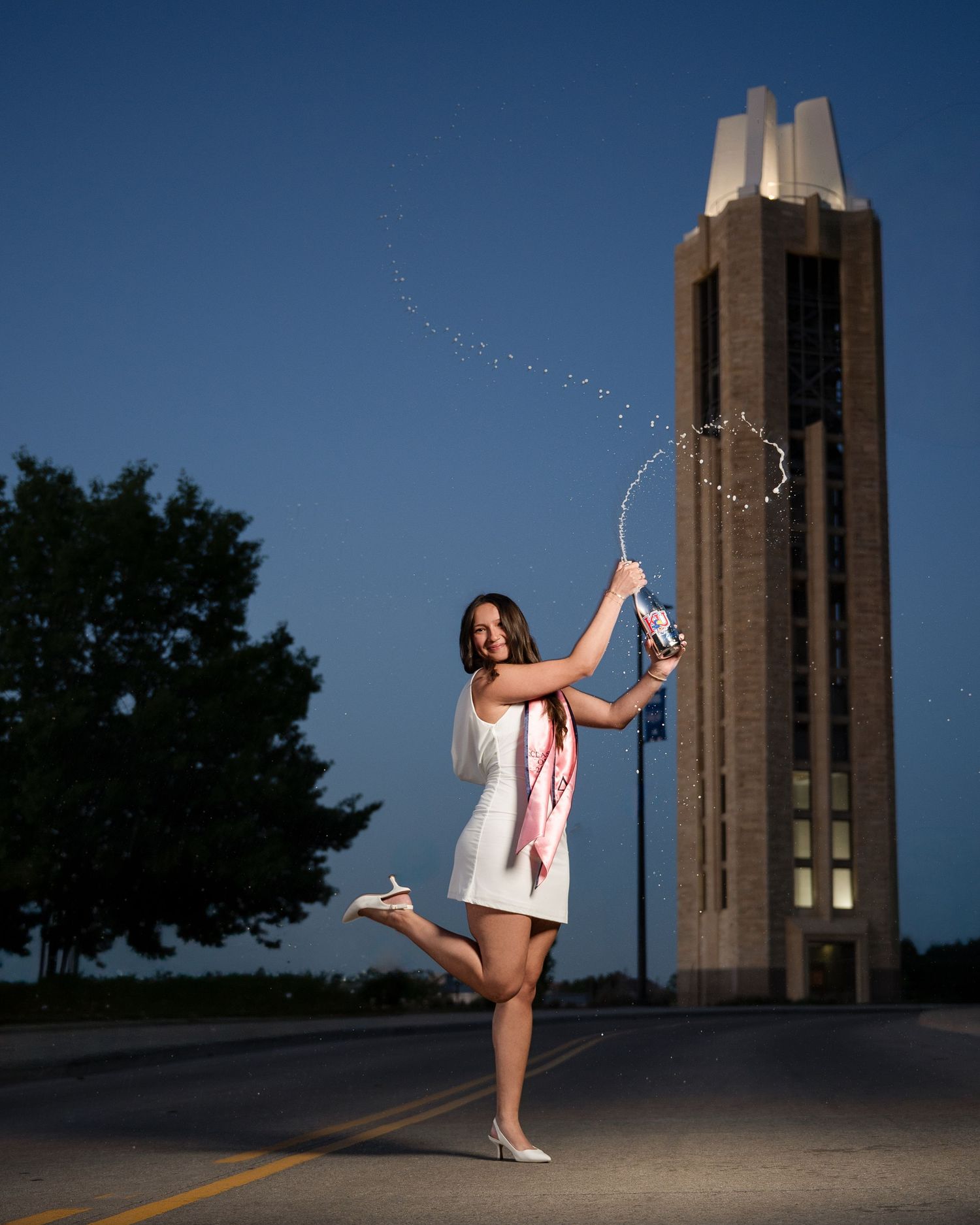 Woman in white dress twirling a light-up hoop near a tall tower at dusk on a quiet road. Kansas City senior pictures Kansas City senior pict
