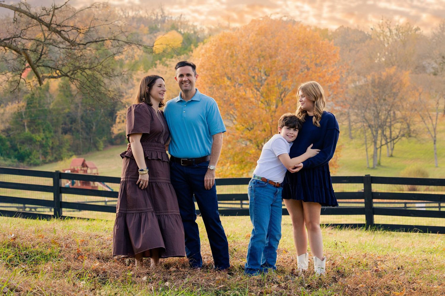 Family of four posing outdoors near a wooden fence with colorful autumn trees and rolling hills in the background.