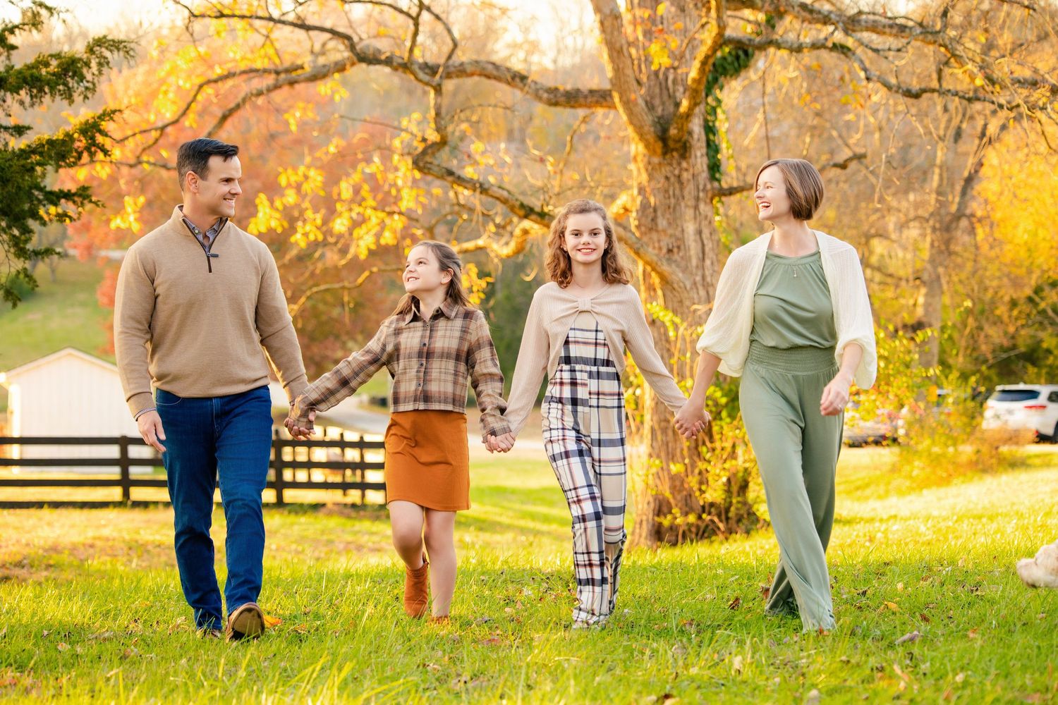 Family of four holding hands walking through a lush green park with colorful autumn trees in background.