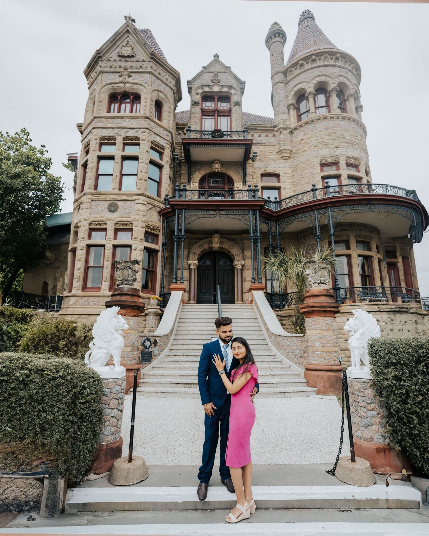 A grand Victorian-era stone mansion with ornate turrets and architectural details features sweeping front steps and landscaping.