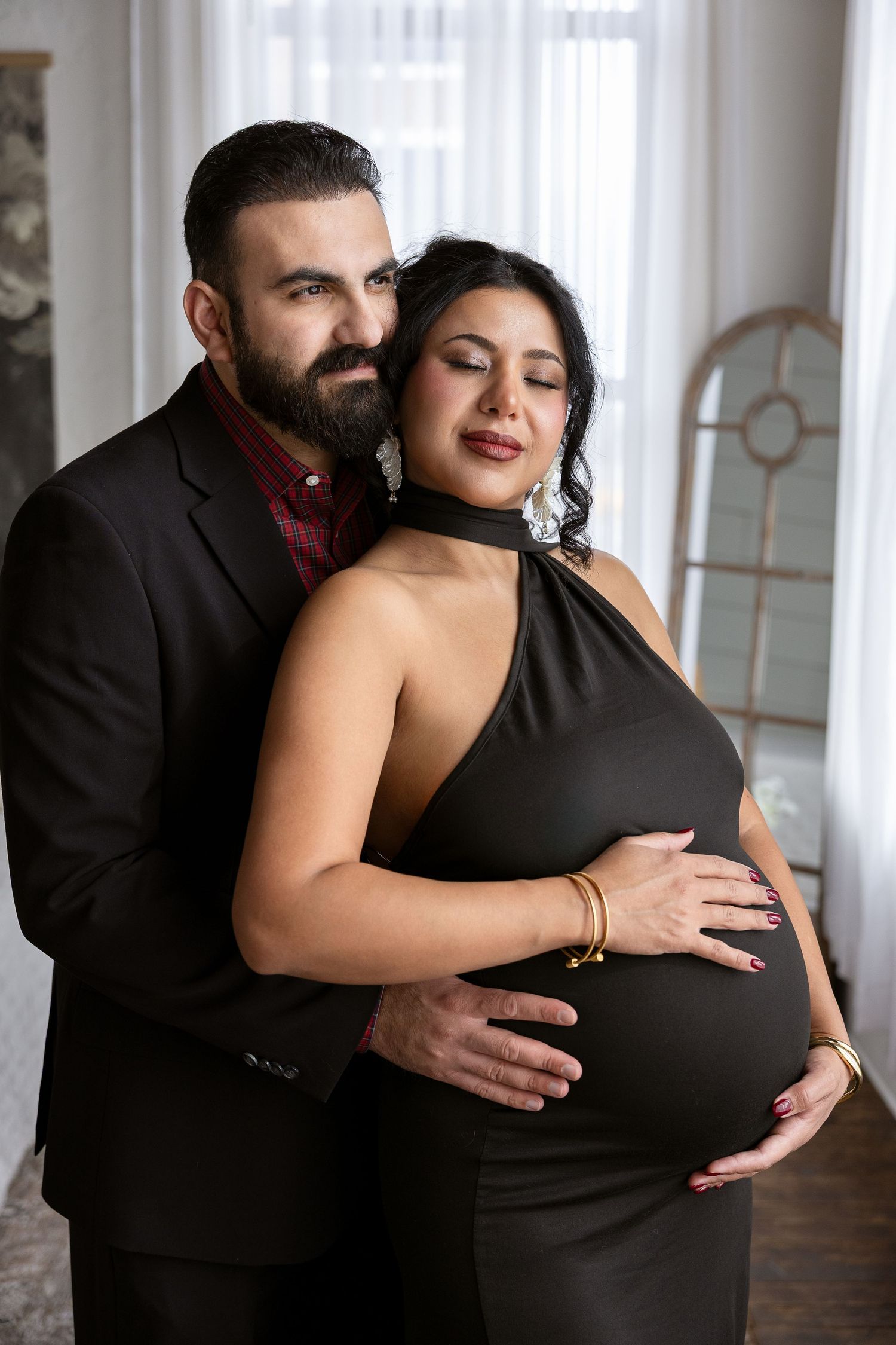 Elegant maternity photo of couple in formal black attire embracing by window with romantic lighting.