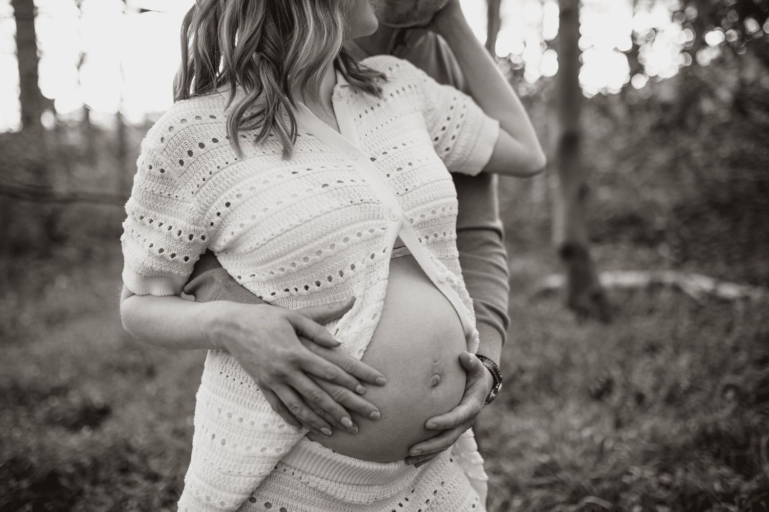 Black and white photo of hands cradling a pregnant belly in a white lace dress outdoors.