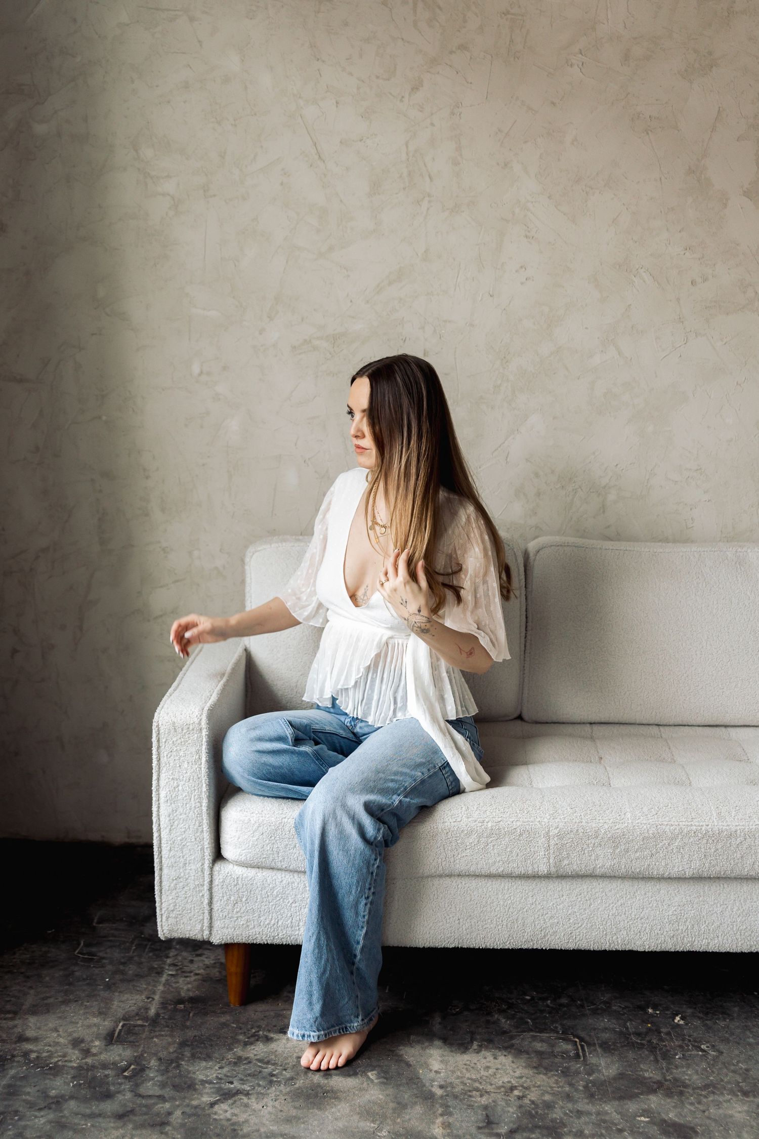 Woman in white blouse and jeans sits casually on light gray couch in minimalist setting against concrete wall.