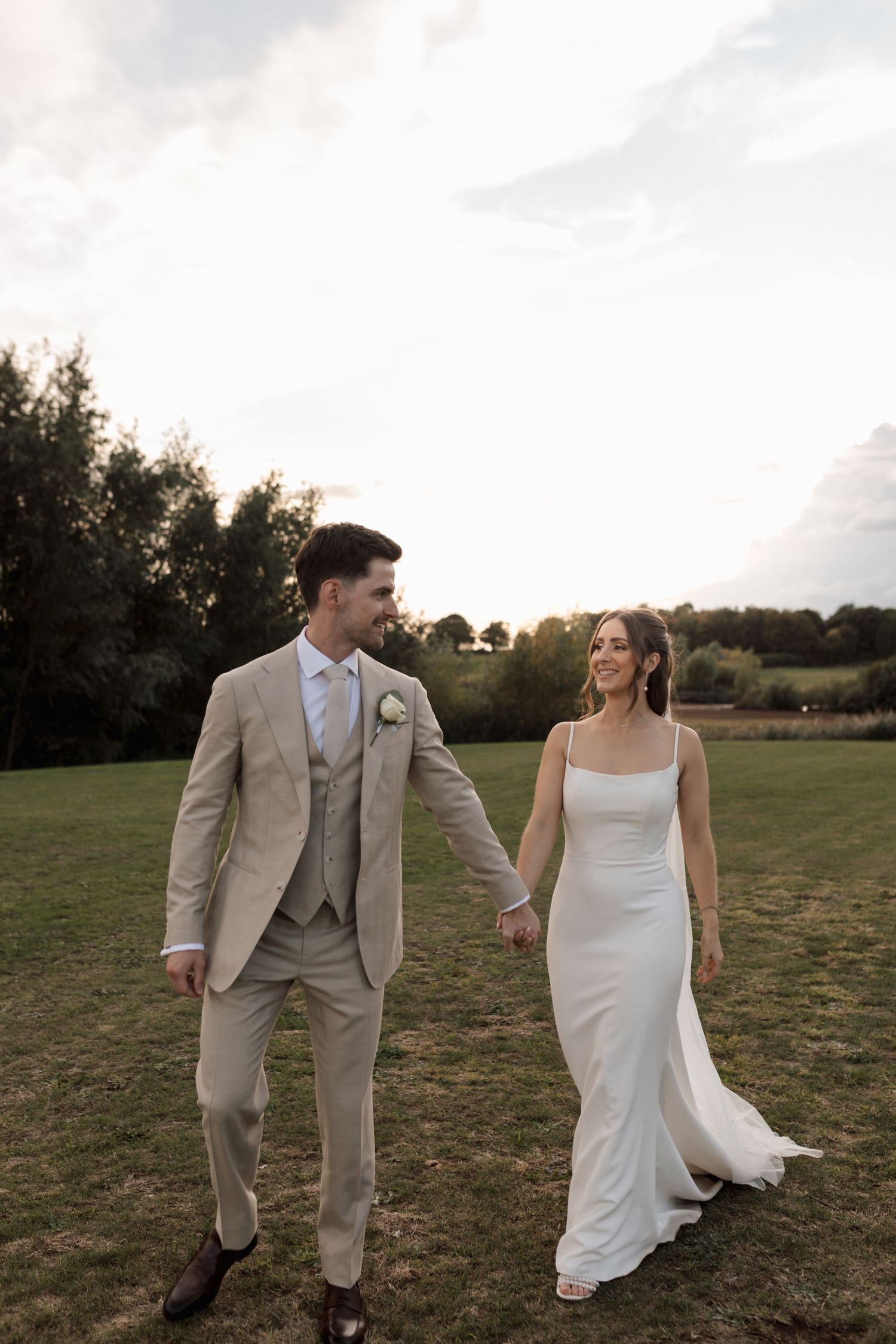 A couple in wedding attire walks hand in hand through a grassy field at sunset.