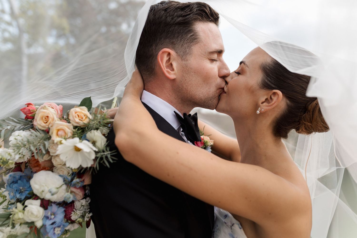 A romantic wedding couple shares a tender kiss while holding a beautiful bouquet of flowers in an outdoor setting.
