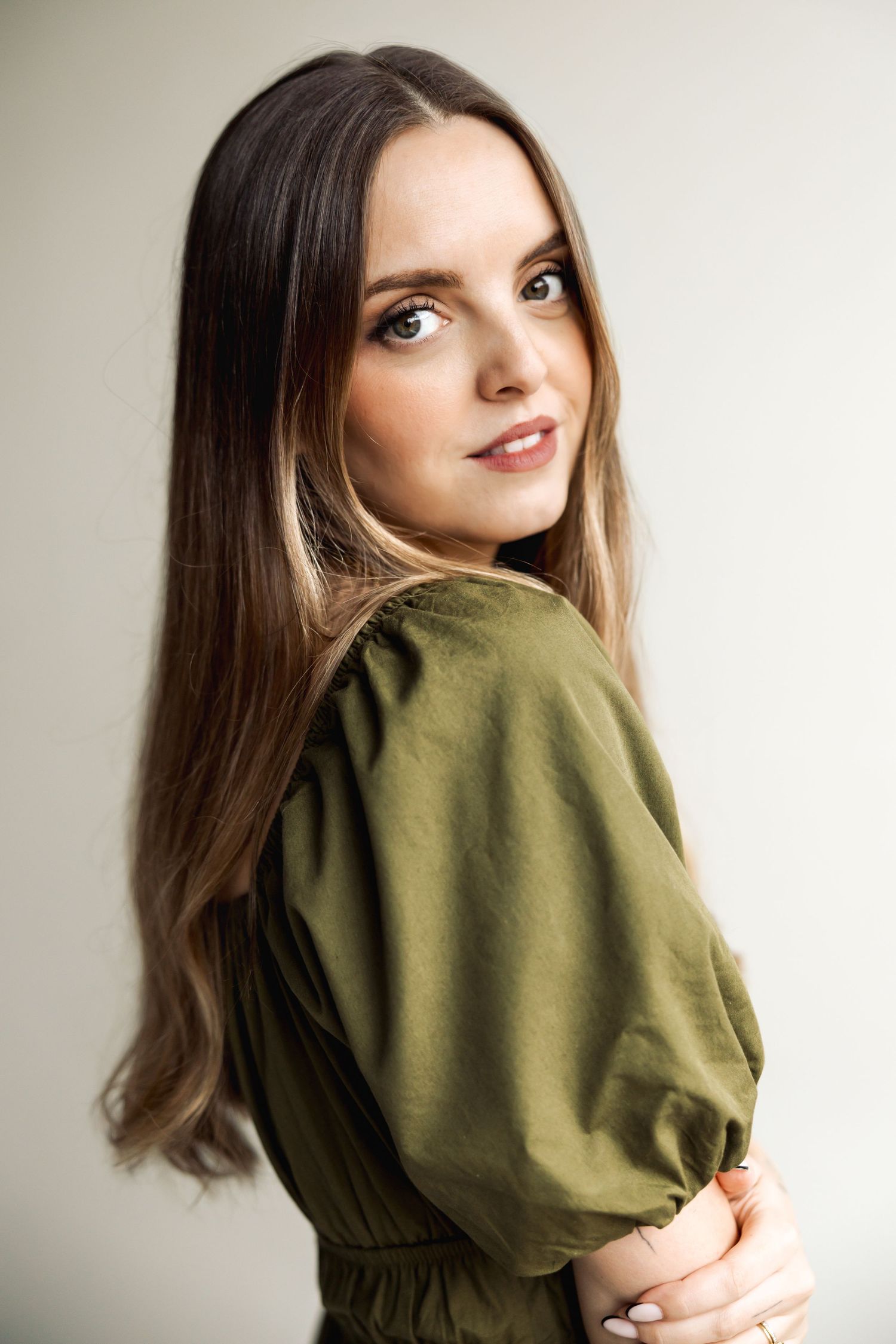 A person with long brown hair poses in a green sweater against a light background.