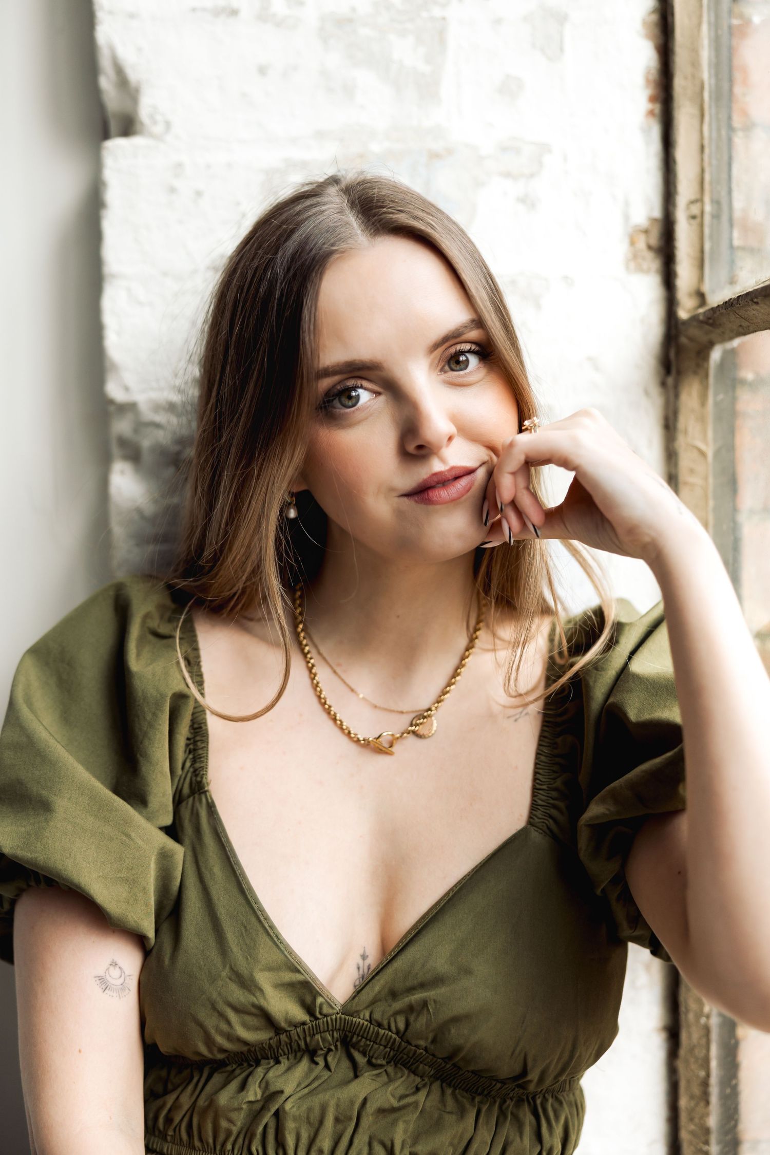 Portrait of someone in an olive green dress with a gold necklace posing thoughtfully against a light wall.