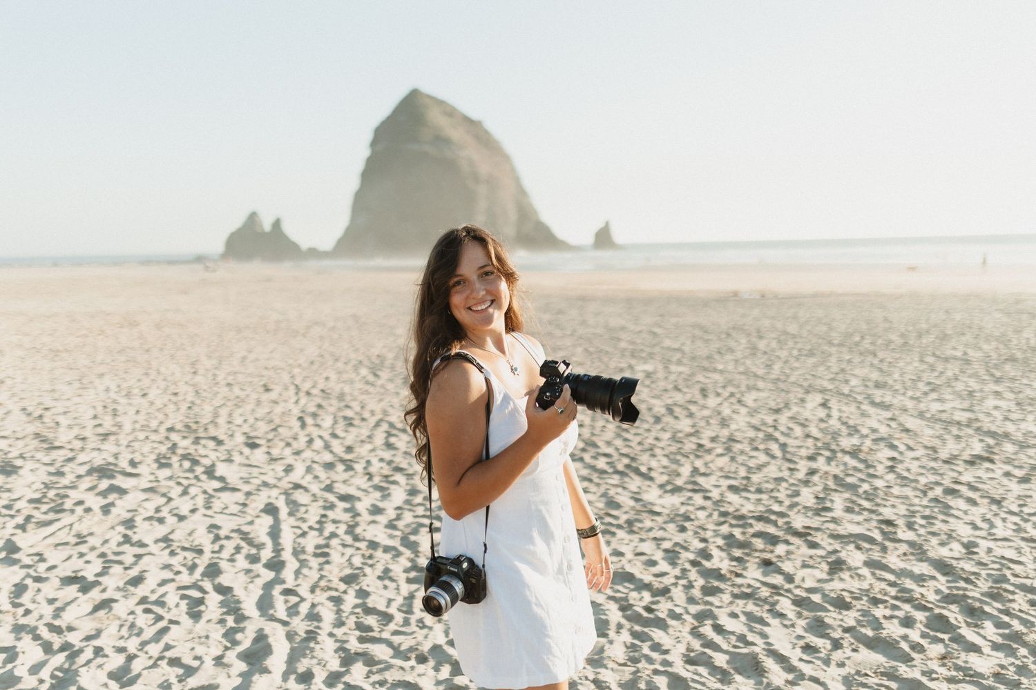 Photographer smiling for photo while holding a camera in front of Haystack Rock at Cannon Beach, Oregon