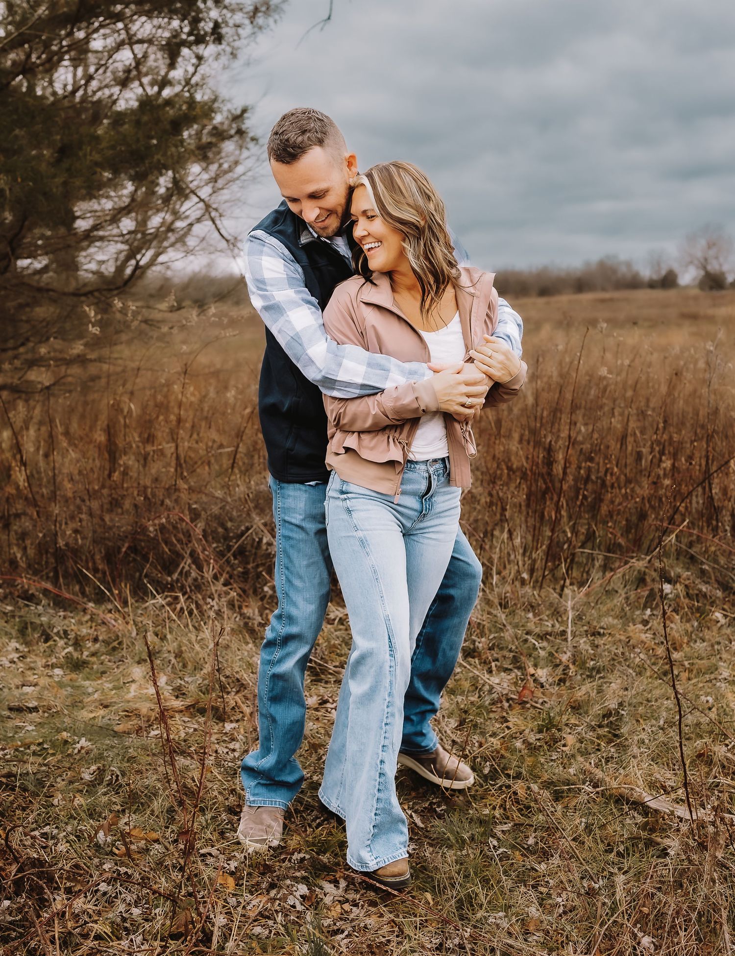 A couple embraces in a rustic field with tall grass and moody skies during their outdoor photo session.