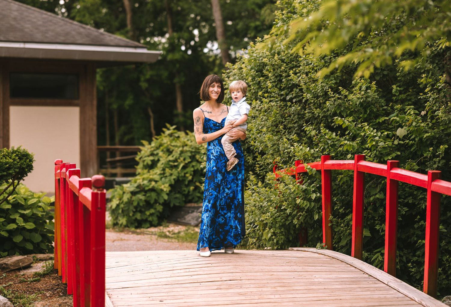 A woman in a blue floral dress stands with a child on a red bridge surrounded by lush green foliage.